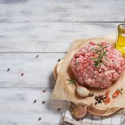 A raw pork sausage on a wooden surface, seasoned with spices and herbs, with a small dab of oil and a backdrop of garlic and spices.