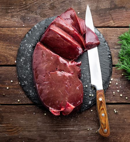 A slice of fresh beef liver on a black cutting board with a knife next to it, surrounded by crushed garlic on the board and scattered herbs in the background.